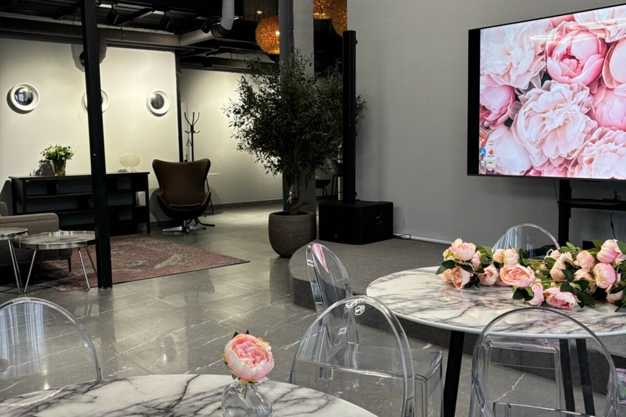 Арендовать Лофт Loftsinoby (Лофтсиноби)  для мероприятий. Современное пространство с серыми стенами и полом. Мраморные столы с прозрачными стульями, цветы в вазах. Кожаное кресло, ковёр, стол, элементы декора и растения. Яркий экран с изображением цветов. Индустриальные детали на потолке.