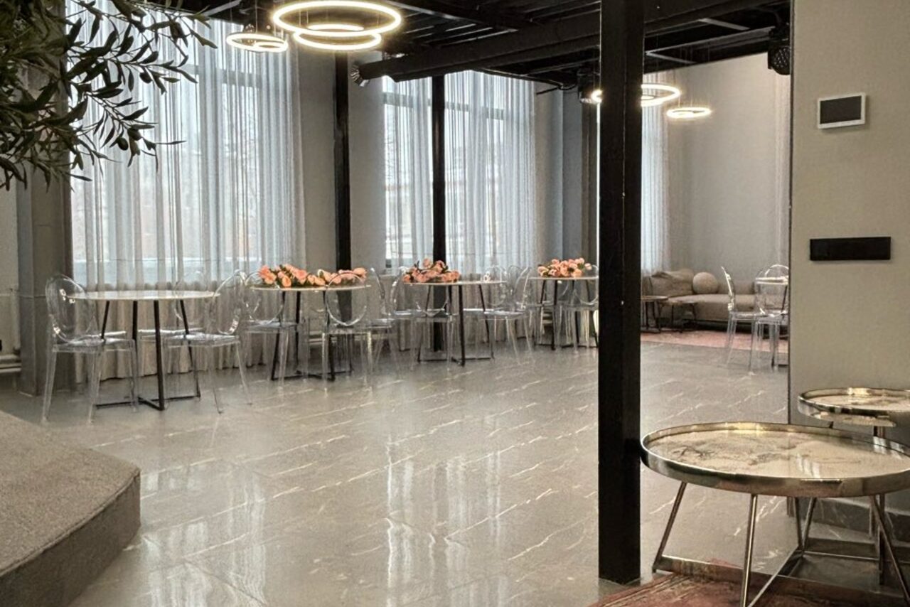 Арендовать Лофт Loftsinoby (Лофтсиноби)  для мероприятий. Просторное помещение с высокими потолками, пол выложен светлой плиткой. Стеклянные столы с прозрачными стульями вдоль стен, неоновое освещение, большие окна с белыми шторами. Стена украшена живой зеленью, есть зона с мягкими диванами.