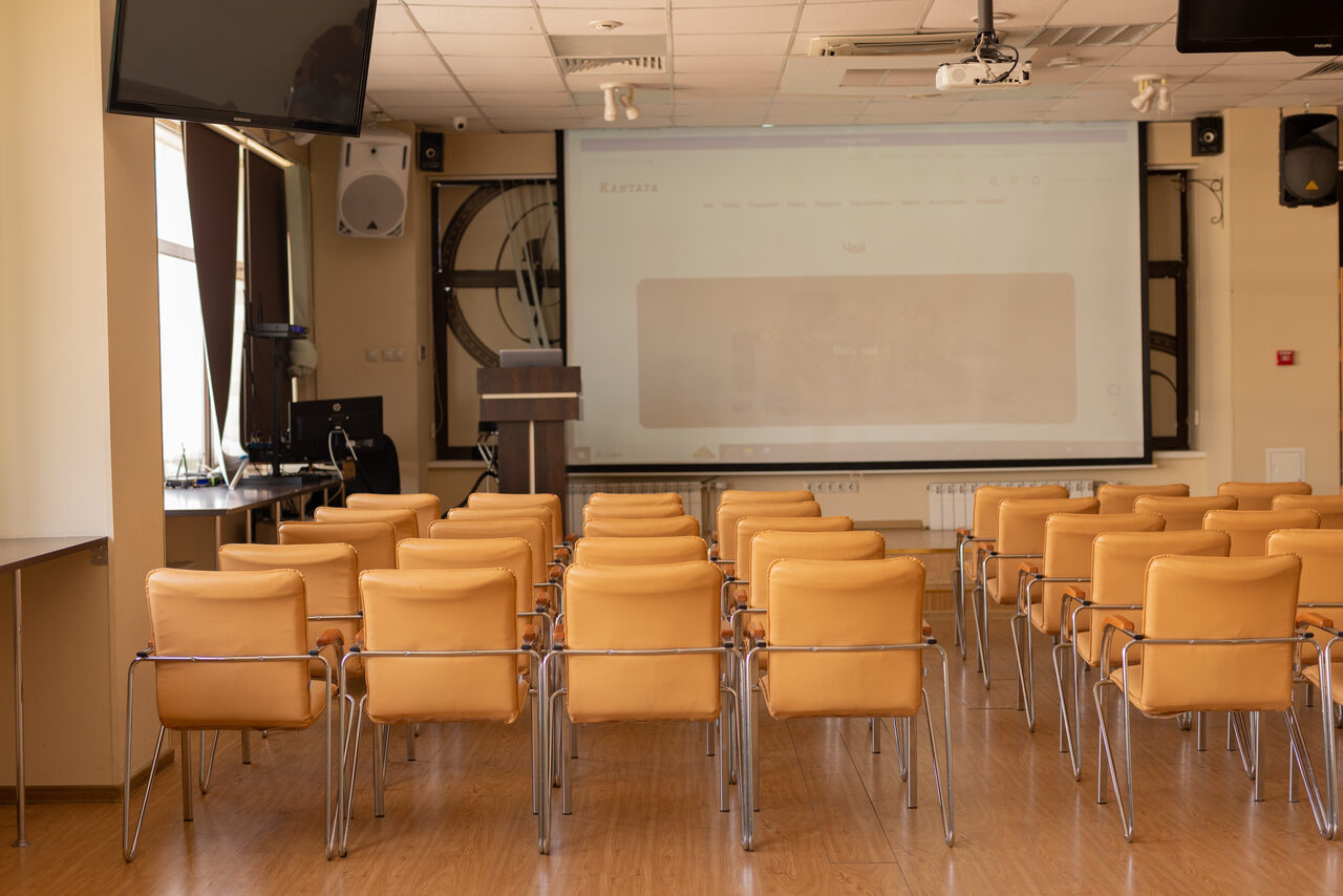 Арендовать Зал 9. Просторный конференц-зал до 200 человек в Академии Кантаты. Конференц-зал с рядами светло-коричневых стульев, деревянный пол, большой экран с проектором, трибунная стойка, светлые стены, потолочные светильники, окна с темными шторами, настенные динамики, кондиционер.