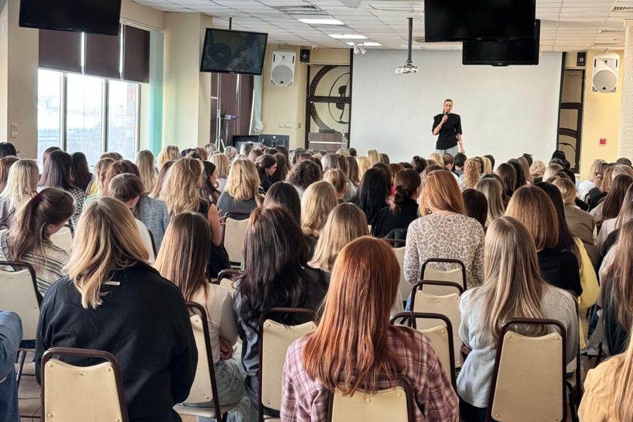 Арендовать Зал 9. Просторный конференц-зал до 200 человек в Академии Кантаты. Конференц-зал с большим количеством людей на стульях, человек в чёрном у белого экрана, микрофоны, две большие колонки на стенах, окна с жалюзи, два телевизора, светлый пол и потолок с люминесцентными лампами.