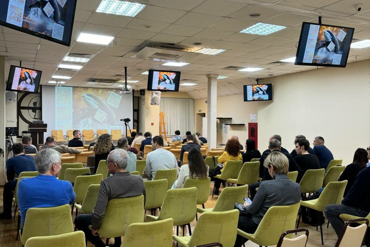 Арендовать Зал 9. Просторный конференц-зал до 200 человек в Академии Кантаты. Конференц-зал с участниками, сидящими на зелёных стульях. Впереди мягкие кресла и проектор, на экранах транслируются изображения. Потолок со встроенным освещением и решетчатыми панелями. На стенах висят экраны. Пол деревянный.