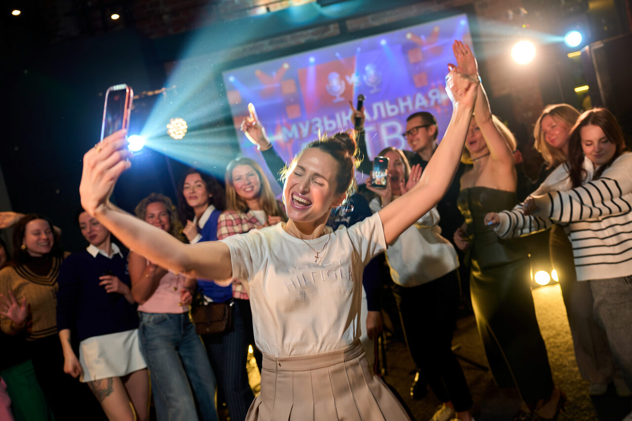 Арендовать Лофт для мероприятий с гольф-симулятором. Человек с телефоном в центре группы людей. Задний план освещён яркими прожекторами, проекция с текстом "Музыкальная" на экране. Несколько человек держат телефоны, создавая эффект мероприятий. Тёмный фон, повседневная одежда.