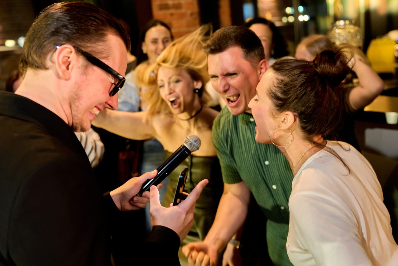 Арендовать Лофт для мероприятий с гольф-симулятором. Изображение показывает группу людей на мероприятии в помещении. Один человек в очках держит микрофон, остальные вокруг в движении, у них радостные выражения лиц. Слева кирпичная стена, видно приглушенное вечернее освещение, барную стойку сзади.