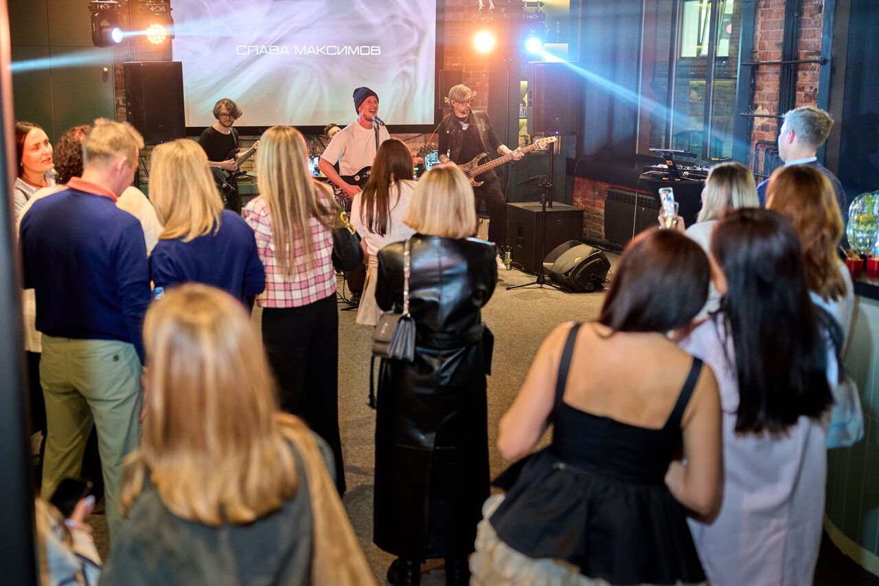 Арендовать Лофт для мероприятий с гольф-симулятором. Музыкальное мероприятие в помещении с кирпичными стенами. Группа выступает на сцене, световое оборудование создает яркое освещение. В центре пространство для стоячей публики. Люди в повседневной одежде, многие снимают видео. На экране над сценой текст.