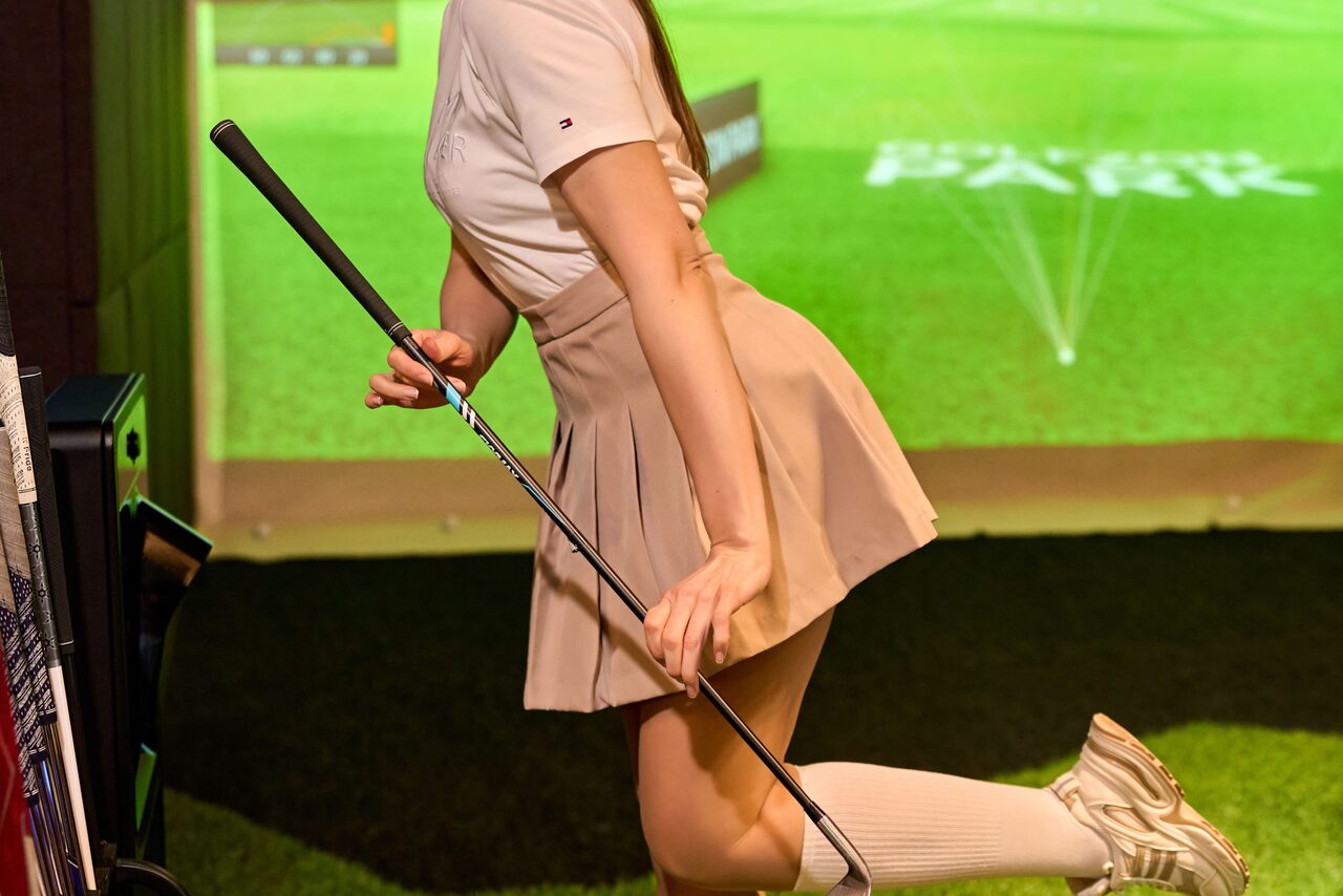 Арендовать Лофт для мероприятий с гольф-симулятором. Изображение человека в светлой одежде с клюшкой для гольфа на фоне зелёного экрана, имитирующего поле для гольфа. Сзади видна проекция с текстом. Пол покрыт искусственной зелёной травой.