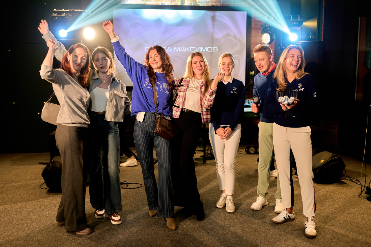 Арендовать Лофт для мероприятий с гольф-симулятором. Просторное помещение с серым ковровым покрытием, сцена с музыкальным оборудованием, проекционный экран с видимыми буквами. Яркое сценическое освещение с несколькими направленными световыми приборами. Группа людей стоит перед экраном.