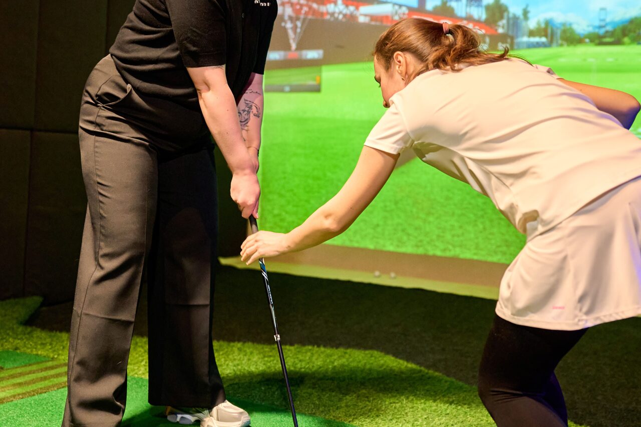 Арендовать Лофт для мероприятий с гольф-симулятором. Интерактивное гольф-пространство с искусственной травой. Два человека взаимодействуют с клюшкой для гольфа. На заднем плане проекционный экран с изображением гольф-поля. Отсутствуют видимые знаки или текст.