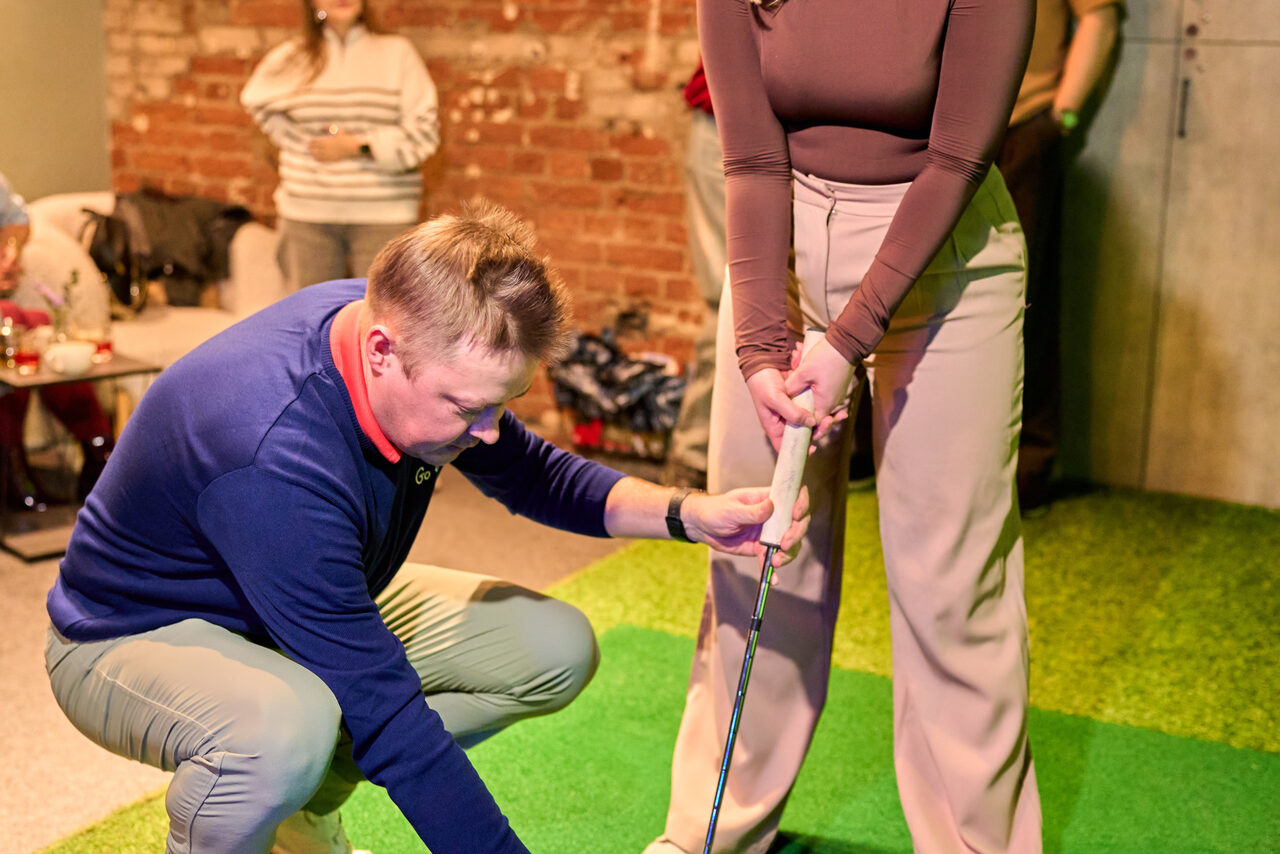 Арендовать Лофт для мероприятий с гольф-симулятором. Человек в синем свитере помогает другому человеку с клюшкой для гольфа на зелёной поверхности, имитирующей траву. На заднем плане кирпичная стена и пара человек, сидящих на бежевых диванах. Яркое освещение.