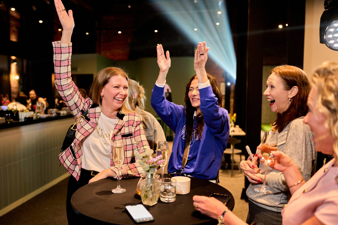 Арендовать Лофт для мероприятий с гольф-симулятором. Барная стойка с высокими круглыми столами, на них стаканы и бокалы, рядом цветы в вазе. Яркое освещение, открытые кирпичные стены, в глубине зеленая стена, темные полы. Потолок со световыми эффектами.