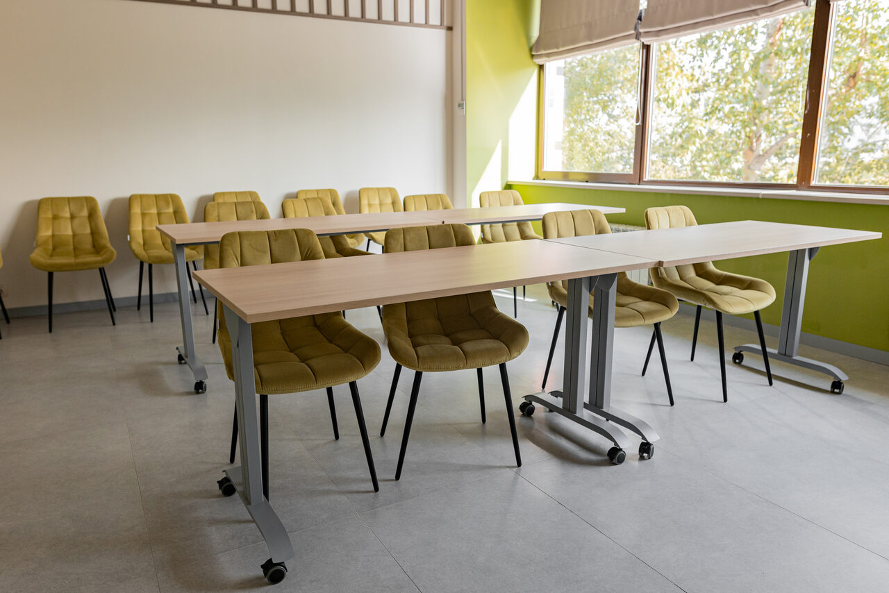 Арендовать Зал 5. Светлый малый конференц-зал до 20 человек в Академии Кантаты. Учебное пространство с двумя столами на колесах и стульями приятного жёлто-зелёного цвета с мягкими сиденьями. Большое окно с бежевой шторой пропускает естественное свет. Пол из светлого материала, стены белые и салатовые.