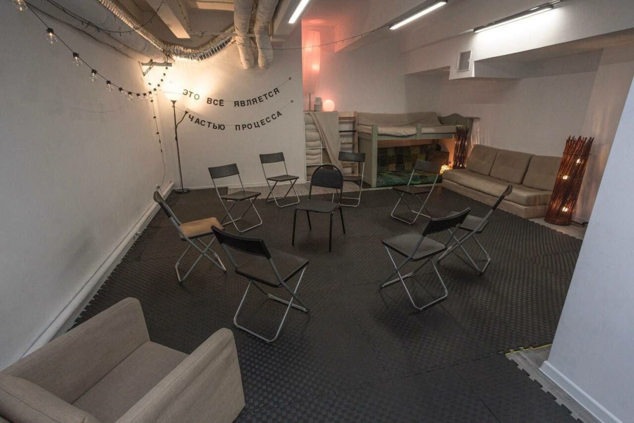 Арендовать Пространство Нора лофт. Комната с черным полом, белыми стенами и мягким освещением. Девять стульев расположены по кругу, слева диван, справа диван и двухъярусная кровать. На стене текст: "ЭТО ВСЕ ЯВЛЯЕТСЯ ЧАСТЬЮ ПРОЦЕССА". Потолок с вентиляционными трубами.