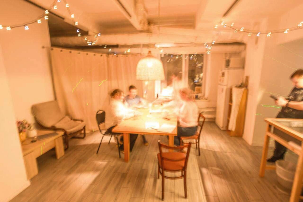 Арендовать Пространство Нора лофт. ```plaintext
Комната с деревянным столом и стульями посередине, мягким креслом слева, холодильником и открытыми полками справа. Светлая плитка на полу, подвесная лампа и гирлянды. Большие окна, зашторенные занавесками. Несколько размытых фигур за столом.
```