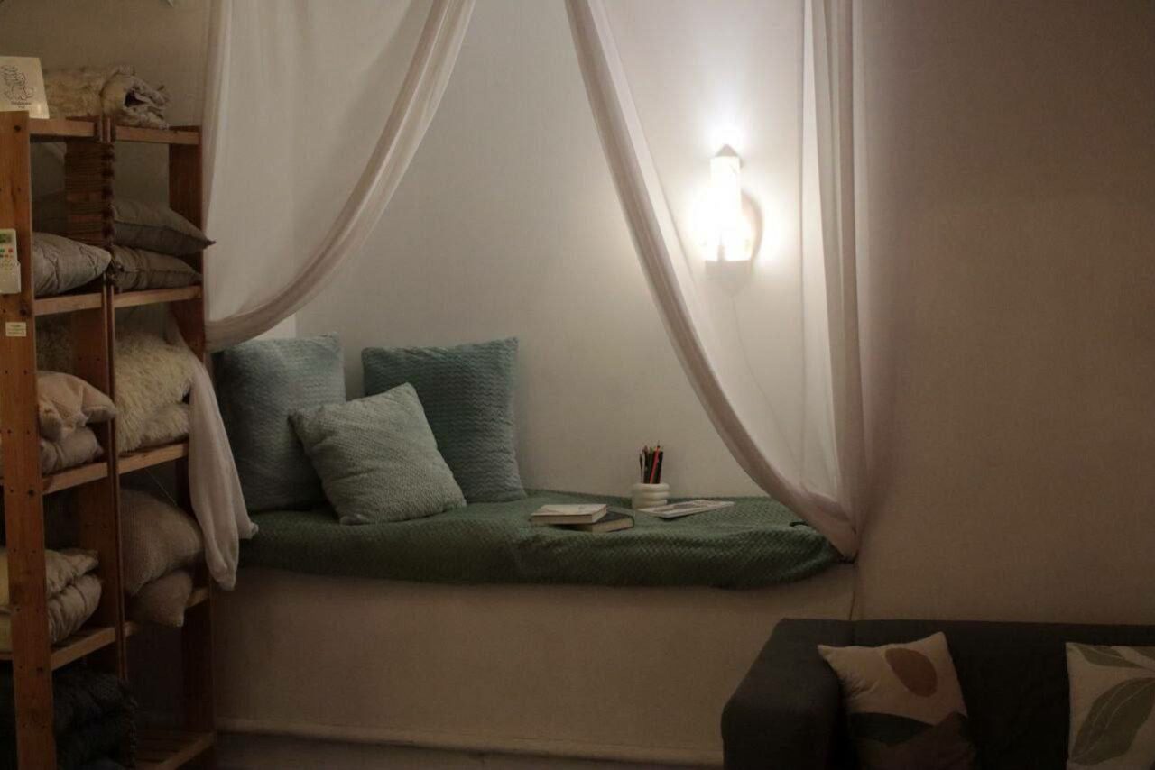 Арендовать Пространство Нора лофт. Пространство с мягкой зоной для отдыха, включающей подушки и книжки, освещённое настенным светильником. Слева деревянные стеллажи с текстилем. Справа диван с подушками. Светлые стены и белый текстиль.