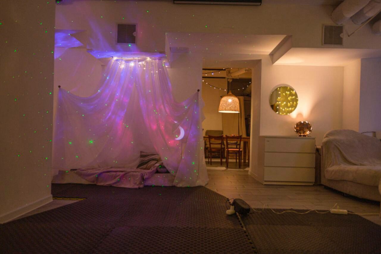 Арендовать Пространство Нора лофт. Комната с зоной отдыха, украшается светящейся занавеской и звездным проектором. Справа комод с круглым зеркалом и лампой. Видны деревянный пол, коврик и стол с деревянными стульями. Мягкое освещение и белые стены.