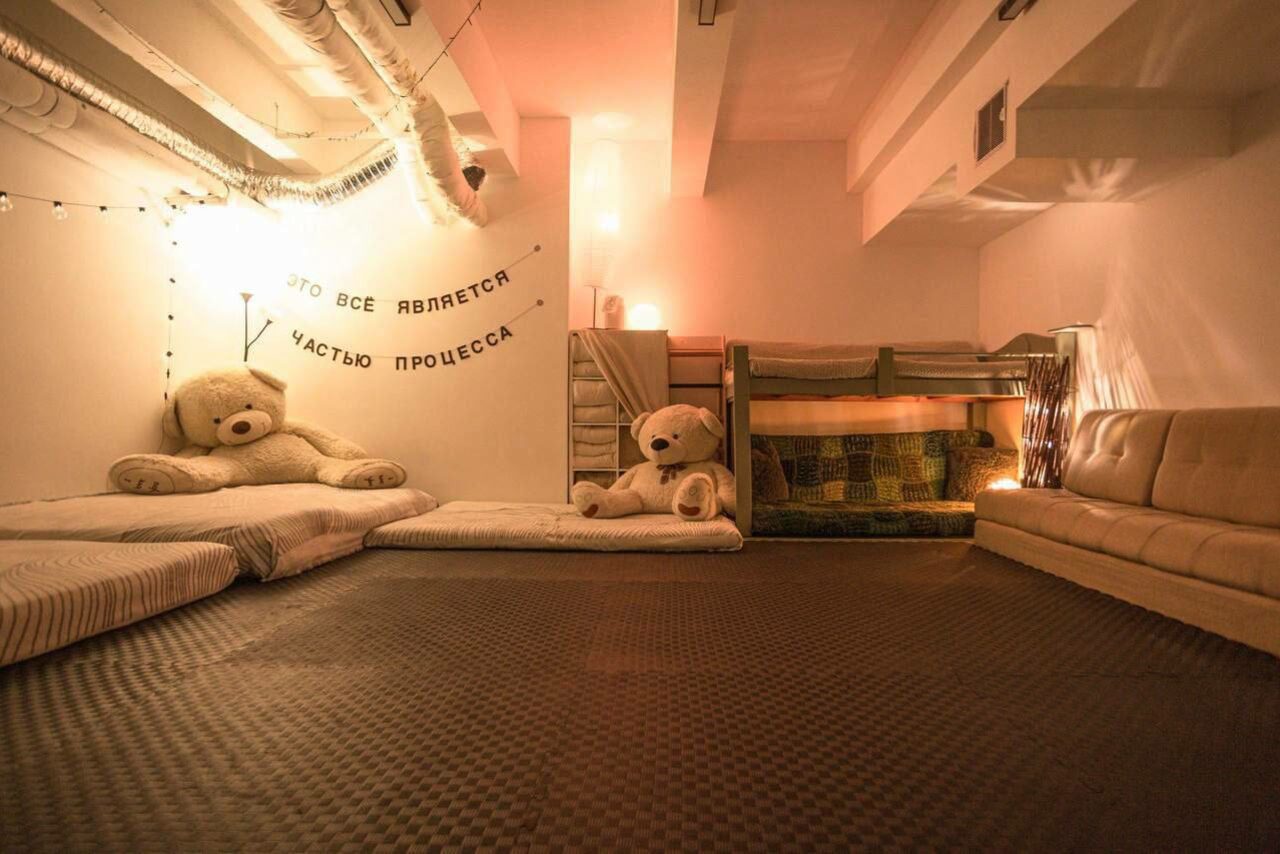 Арендовать Пространство Нора лофт. Подвальное пространство с низкими потолками, ковровое покрытие на полу. На стене текст: "Это все является частью процесса". У стены больших мягких мишек, матрасы, двухъярусная кровать, диван. Мягкое освещение, светодиодные гирлянды.
