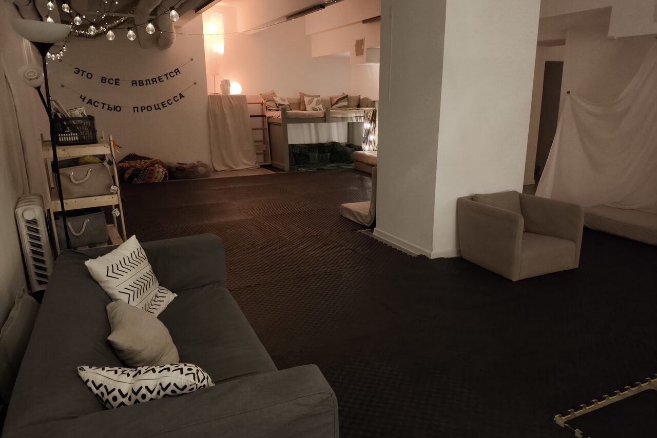 Арендовать Пространство Нора лофт. Лофт-пространство с диванами и креслом серого цвета. Есть полка, светильники и текст на стене. Пол тёмный, стены светлые, освещение мягкое. На полу лежат подушки, на стенах висят светильники и гирлянды.