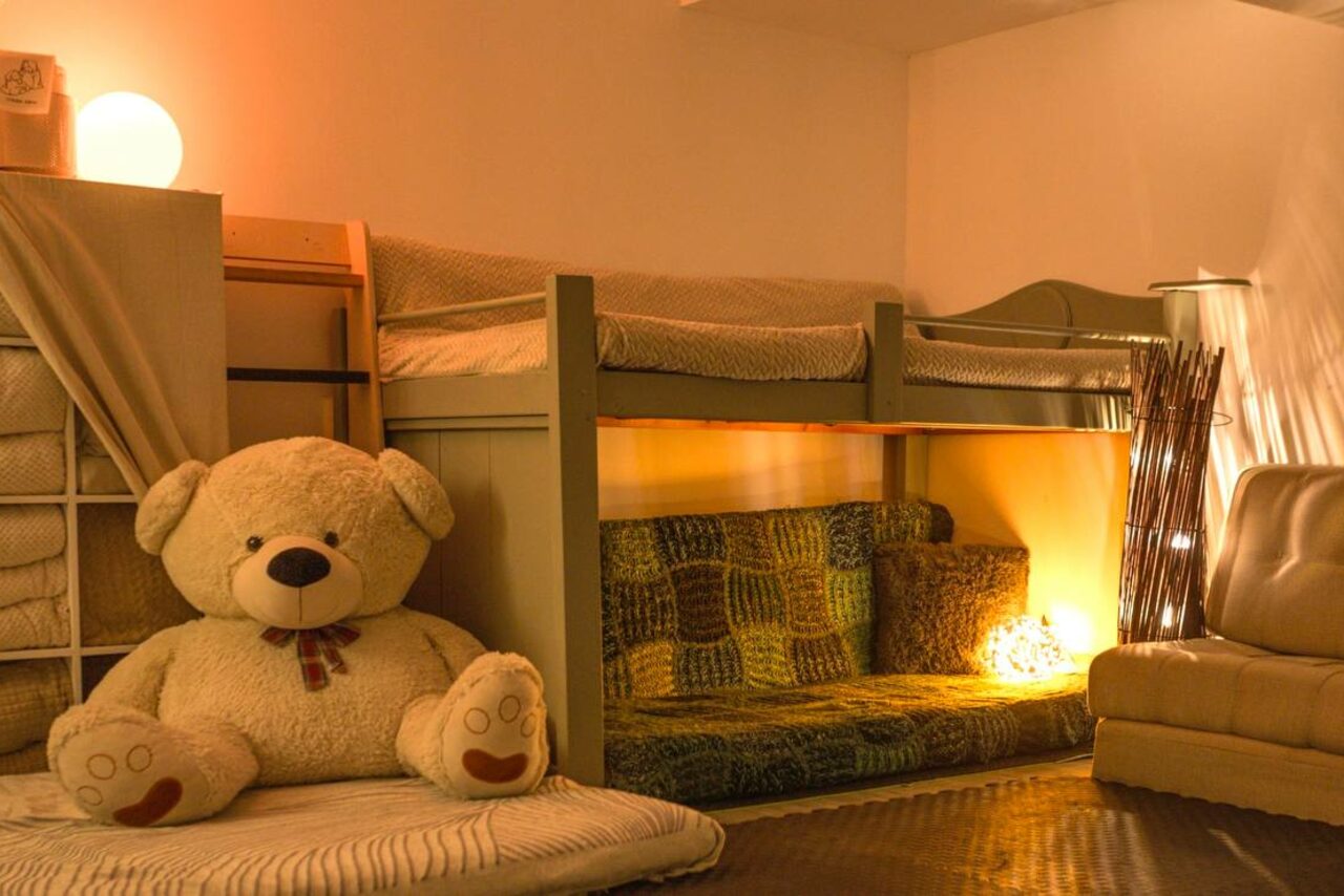 Арендовать Пространство Нора лофт. Двухъярусная кровать с постельным бельём, кресло, большой плюшевый медведь, мягкий диван, тёплое освещение, светильники, пледы на диване, подушки, деревянные элементы интерьера, светло-коричневые и бежевые цвета, спокойная атмосфера.