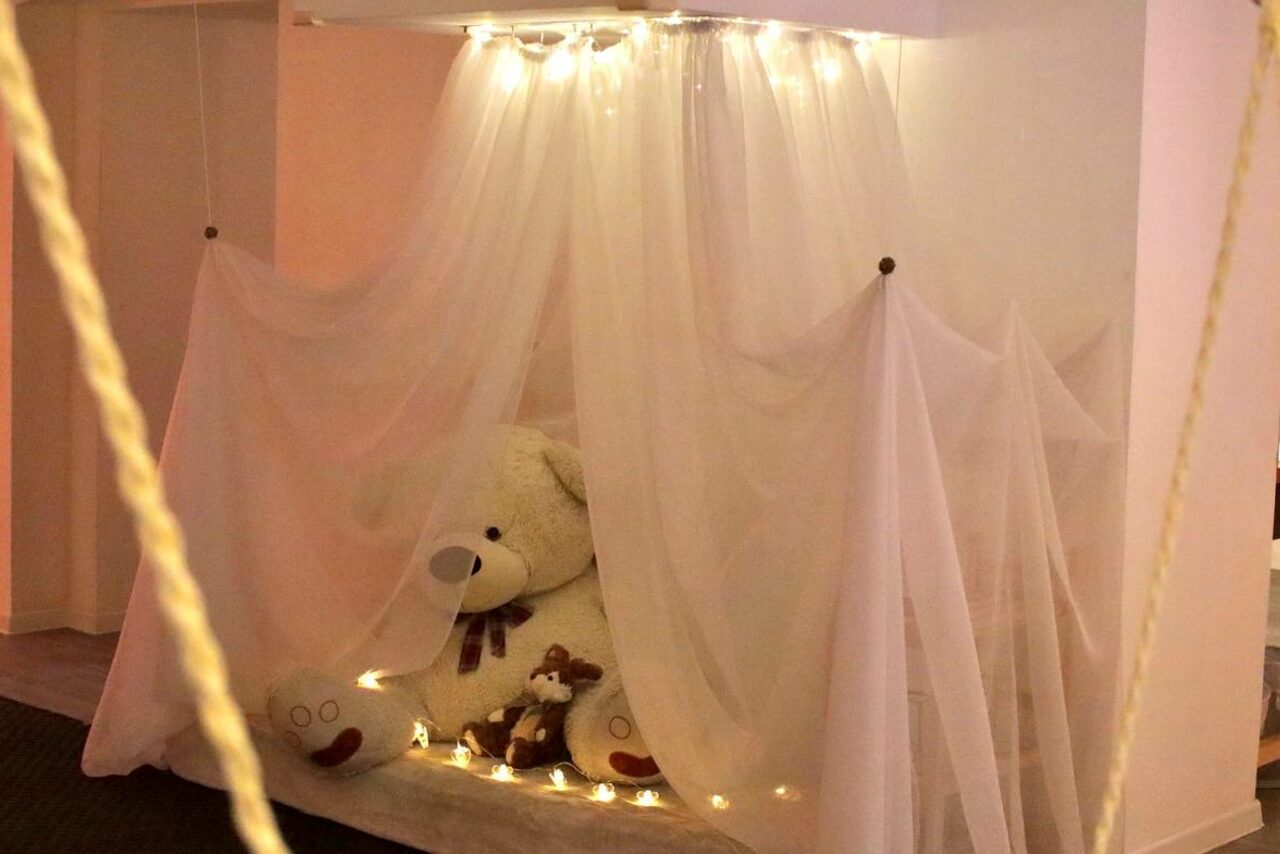 Арендовать Пространство Нора лофт. Уютное пространство с белыми полупрозрачными занавесями, украшенными гирляндами. Внутри большой плюшевый медведь и подушки. Пол светлый, стены белые. Освещение теплое, мягкое. Видны веревочные элементы.