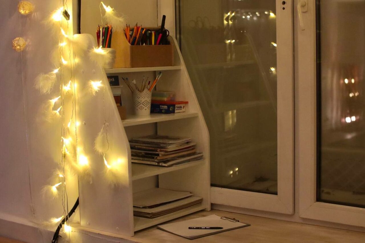 Арендовать Пространство Нора лофт. Настольная зона у окна с полками, заполненными книгами и канцтоварами. На полке стоят карандаши в стаканах. На стене гирлянда с тёплым белым освещением. На столе блокнот с ручкой. Белые рамы окна.