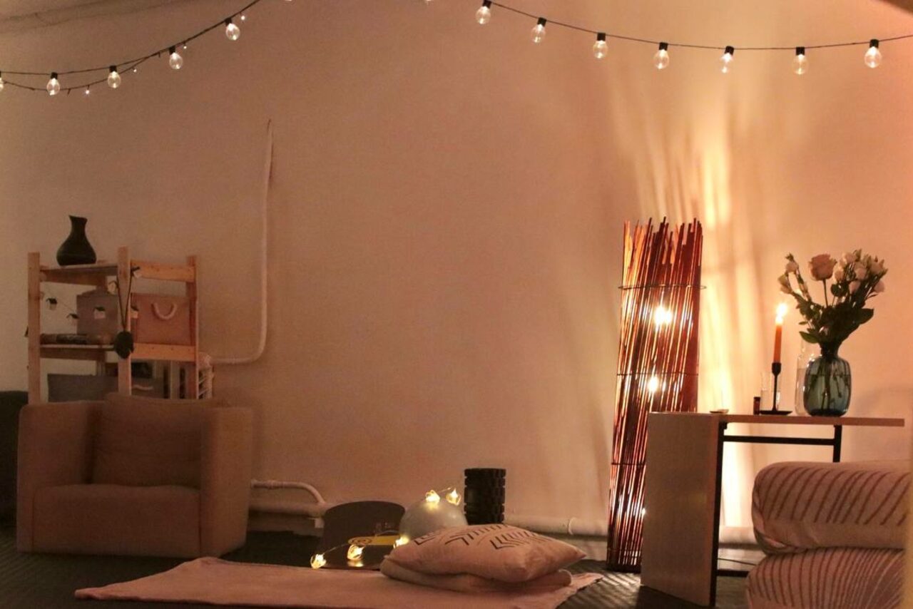 Арендовать Пространство Нора лофт. Уютное пространство с креслом, подушками и пуфом. На стене гирлянда с лампочками, сбоку светильник с ветками. На столике ваза с цветами и свечи. В углу полка с предметами. Мягкое освещение, нейтральные тона.