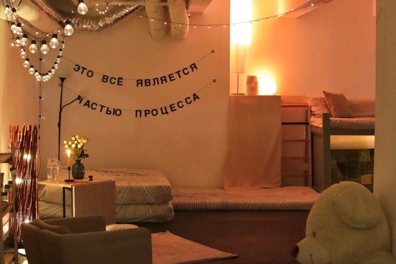 Арендовать Пространство Нора лофт. Уютное пространство с нейтральной цветовой гаммой. Низкие матрасы, кресло, большой плюшевый медведь. На стене надпись: "ЭТО ВСЕ ЯВЛЯЕТСЯ ЧАСТЬЮ ПРОЦЕССА". Мягкое освещение, гирлянды, свечи и вазы с цветами.