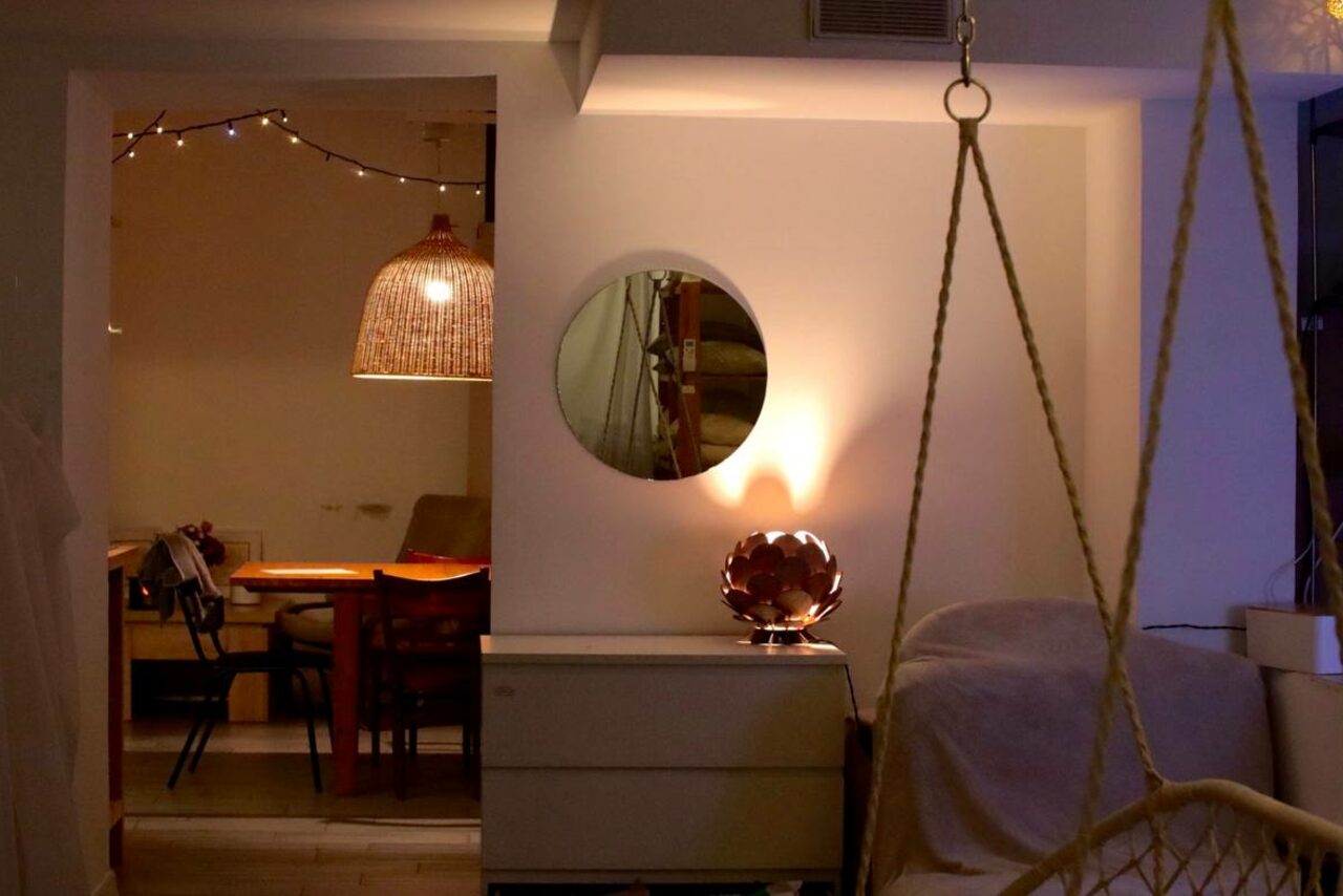 Арендовать Пространство Нора лофт. Современное помещение с обеденной зоной и качелями. Обеденный стол с деревянными стульями, над столом плетеный светильник. Белая стена с круглым зеркалом и настольной лампой. Комфортное освещение, гирлянды, деревянные и текстильные элементы.