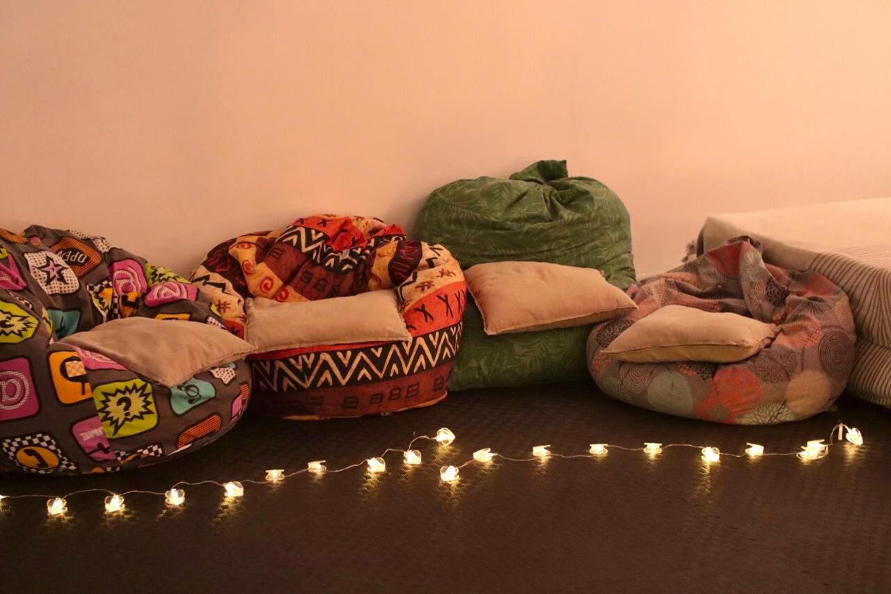 Арендовать Пространство Нора лофт. Мягкое пространство с разноцветными креслами-мешками на тёмном полу, украшенное светодиодной гирляндой. Стены бежевые. На креслах лежат бежевые подушки. Справа виден край дивана с полосатым покрывалом.