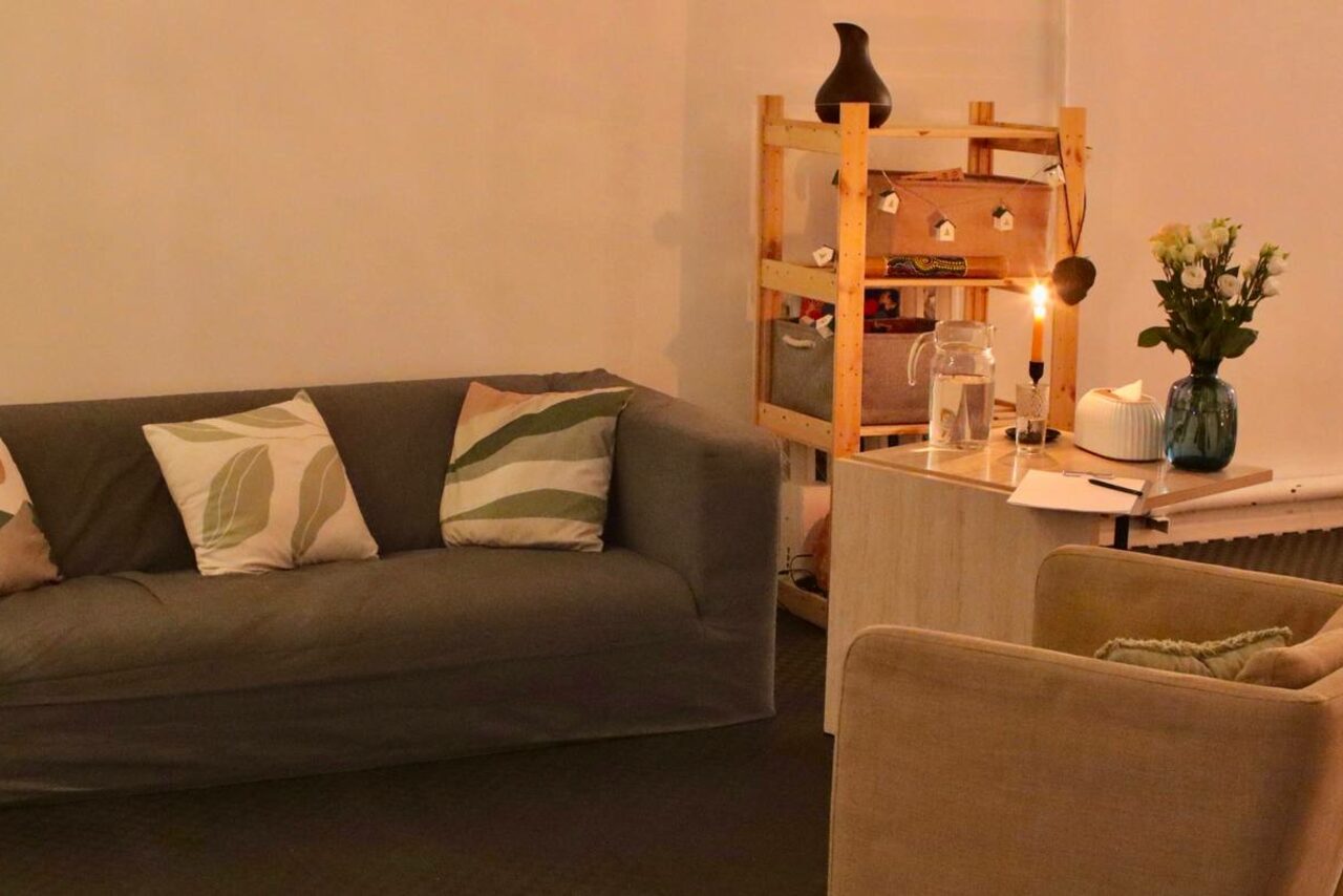 Арендовать Пространство Нора лофт. Гостиная с серым диваном и бежевым креслом, украшенными декоративными подушками. Деревянный стеллаж с декором, лампа с теплым освещением, стол с цветами. Нейтральные бежевые и серые тона, ковровое покрытие.