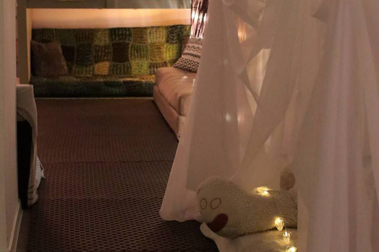 Арендовать Пространство Нора лофт. Комната с темным ковровым покрытием, мягкой мебелью, подушками и текстильным декором в углу. Освещение теплое, рассеянное, украшено светодиодными гирляндами. Стены светлые, текстиль белый и зеленый.