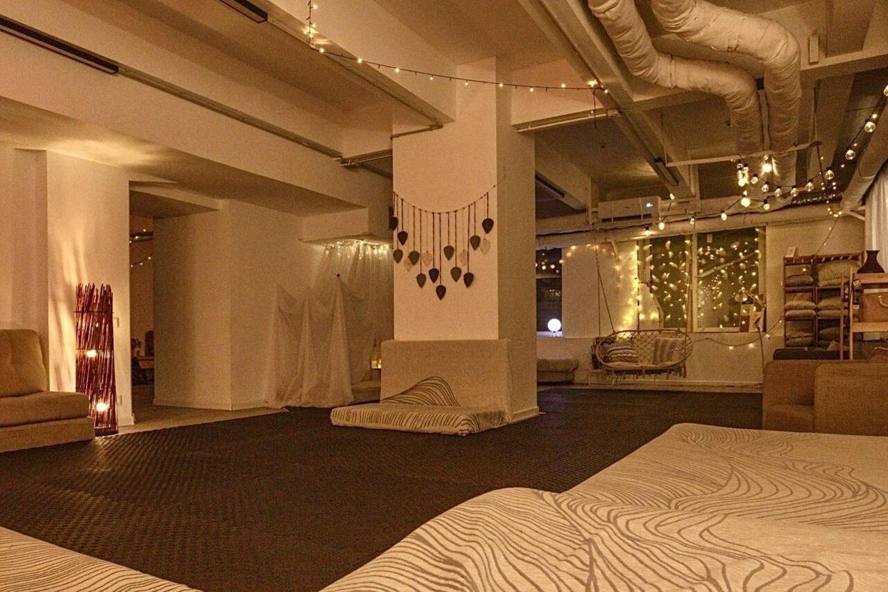 Арендовать Пространство Нора лофт. Просторное помещение с белыми стенами и потолком. На полу тёмные ковры. Мебель: мягкие диваны, пуфы. Декор из гирлянд и текстильных украшений. Потолок с видимыми вентиляционными трубами. Освещение мягкое, от гирлянд и настольных ламп.