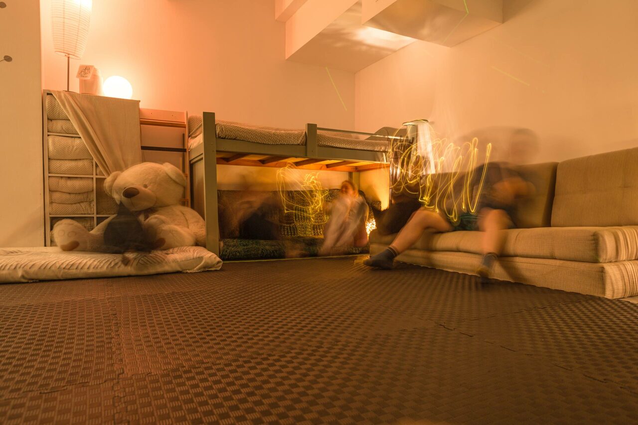 Арендовать Пространство Нора лофт. Комната с двухъярусной кроватью, расположенной у стены. На полу ковровое покрытие темного цвета. Рядом с кроватью большой плюшевый медведь и мягкий диван. Освещение теплое, лампа на полу создаёт мягкий свет. Стены светлые.