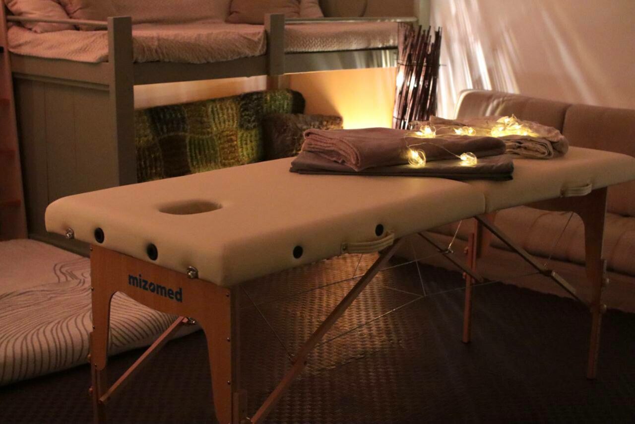 Арендовать Пространство Нора лофт. Массажный стол с полотенцами, украшенными гирляндой, стоит перед диваном. На заднем плане двухъярусная кровать с матрасами и подушками. Освещение мягкое, тёплое; на полу плитка тёмного цвета.