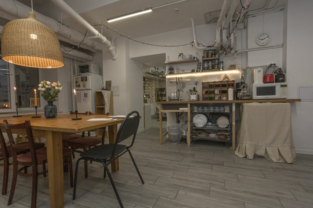 Арендовать Пространство Нора лофт. Кухонное пространство с деревянным обеденным столом и разными стульями, открытые полки с посудой и оборудованием, подвесная плетеная лампа, настенные часы, белые стены, светлая плитка на полу, вентиляционные трубы на потолке, свечи и цветы на столе.