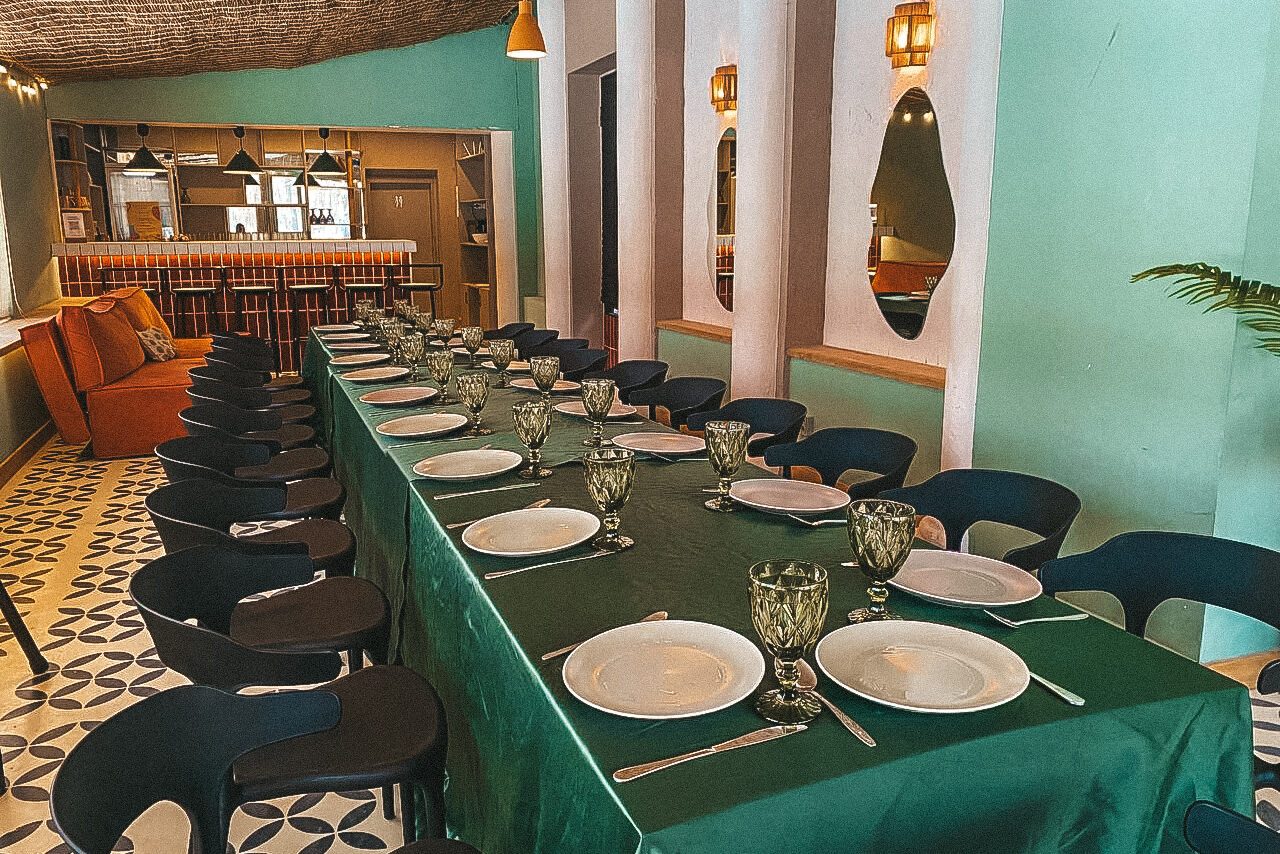 Арендовать Уникальное пространство для мероприятий в Сокольниках. Пространство с длинным столом, покрытым зеленой скатертью, окруженным черными стульями. На столе белая посуда и стеклянные бокалы. Бежевые и зеленые стены, овальные зеркала, подвесные светильники, декоративная плитка на полу, оранжевый диван.