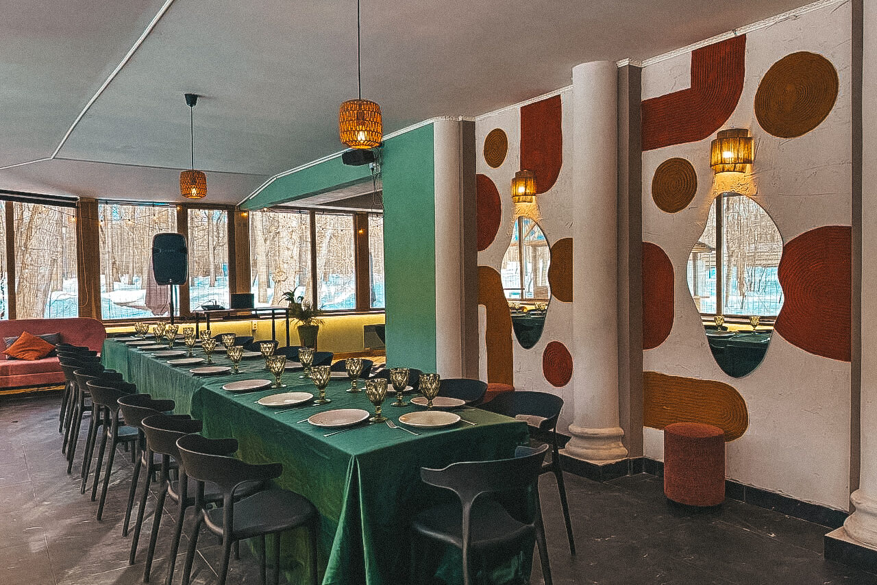 Арендовать Лофт с панорамными окнами в парке Сокольники. Просторная обеденная зона с длинным столом, покрытым зеленой скатертью; черные стулья; на столе стоят бокалы и тарелки. На стенах декоративные круги разных цветов. Большие окна дают естественный свет. Потолочные светильники и колонки у окна.