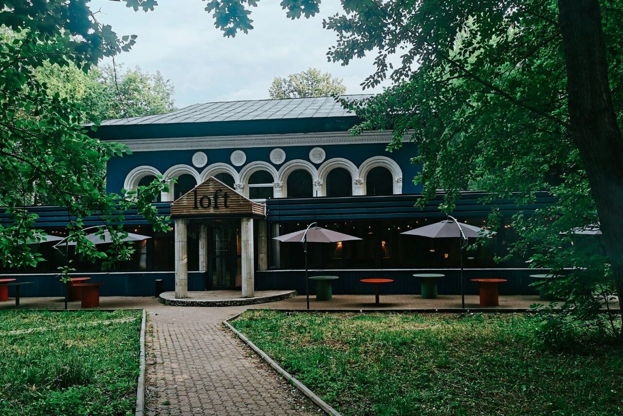 Арендовать Лофт с панорамными окнами в парке Сокольники. Двухэтажное здание с большими арками и тёмным фасадом, окруженное деревьями. Перед зданием расположены столы с зонтиками и кашпо с растениями. Дорожка из плитки ведет к входу с деревянной крышей и надписью "loft".