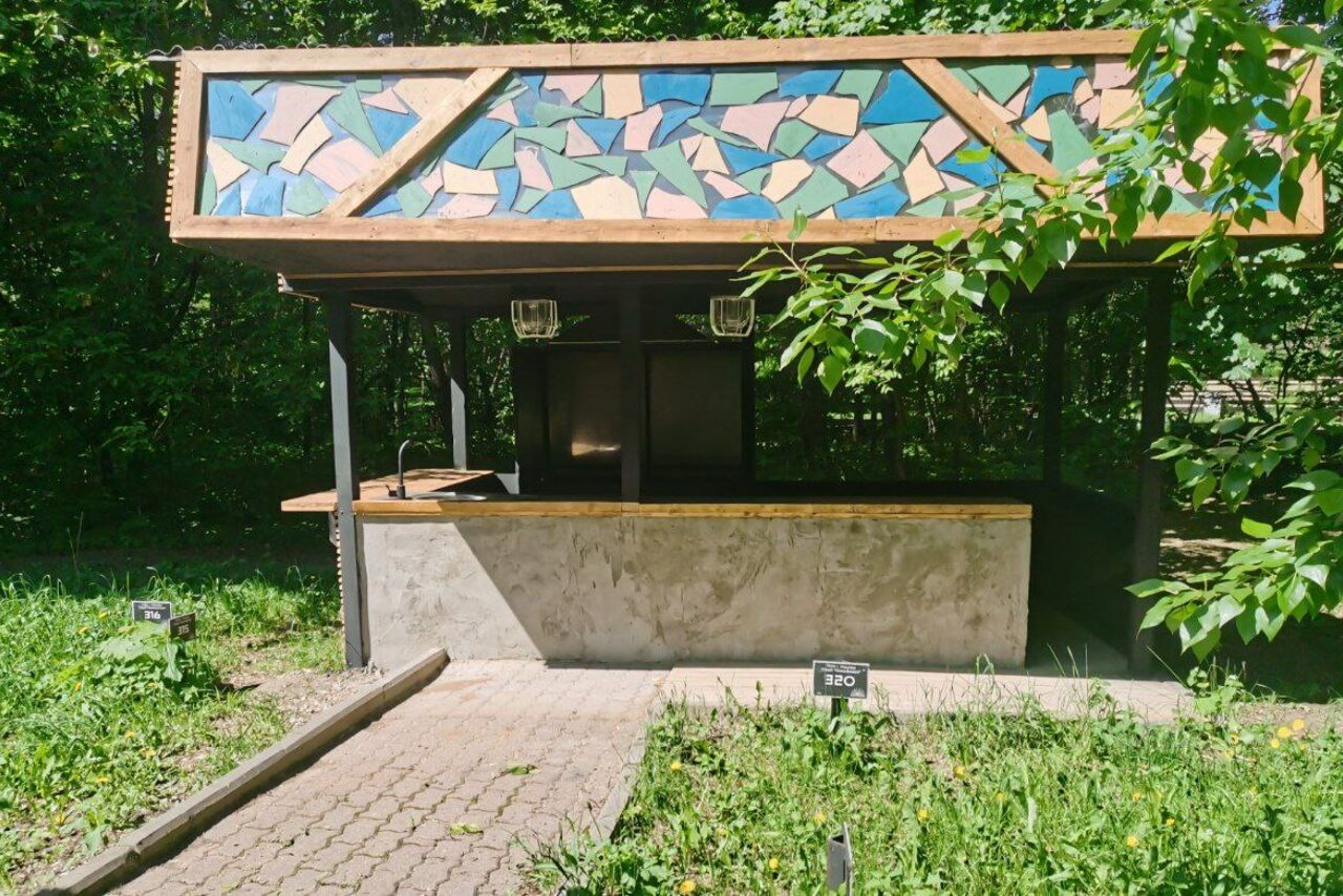 Арендовать Лофт с панорамными окнами в парке Сокольники. Павильон с бетонной стойкой, крыша украшена мозаикой из цветного стекла. Плиточная дорожка ведет к павильону сквозь траву. Внутри два металлических светильника. Вокруг деревья и зелень. Видны небольшие таблички.