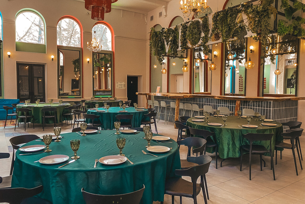 Арендовать Просторный и светлый лофт с панорамным видом в парк. Просторное помещение с круглыми столами, покрытыми зелеными скатертями, и черными стульями. На стене крупные зеркала, украшенные зеленью и светильниками. Высокие окна с арочными проемами, на потолке люстры. Светлые стены и пол.