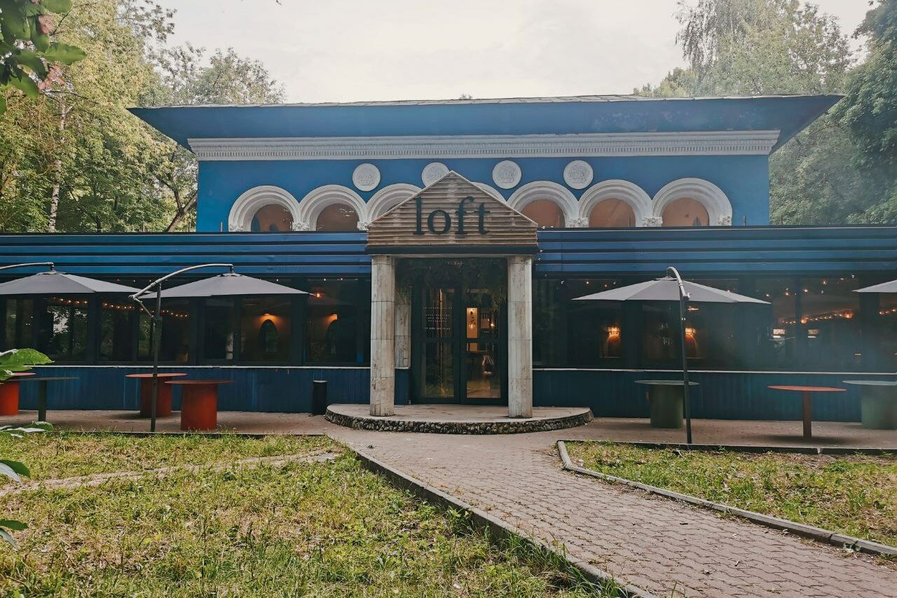 Арендовать Просторный и светлый лофт с панорамным видом в парк. Здание синих тонов с большими арочными окнами, центральный вход с деревянной вывеской "loft", дорожка из плитки ведет к входу, вокруг зеленая трава, столы с красными и зелеными ножками под серыми зонтами.