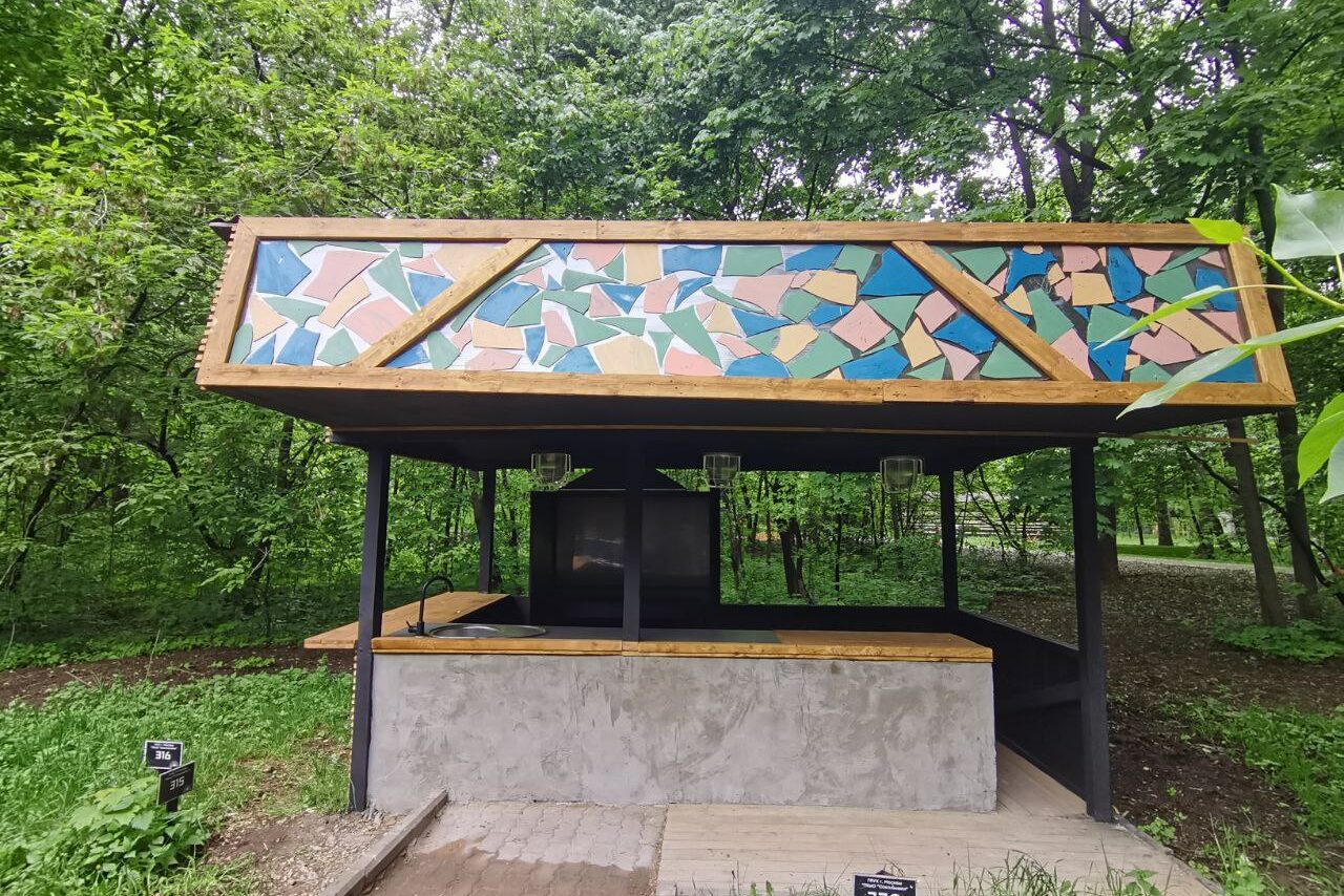 Арендовать Просторный и светлый лофт с панорамным видом в парк. Крытое открытое пространство с бетонной стойкой, тёмной металлической конструкцией и пёстрой мозаичной панелью на крыше, состоящей из разноцветных элементов. Освещение из подвесных светильников. Окружено зеленью леса. Дорожка из брусчатки.