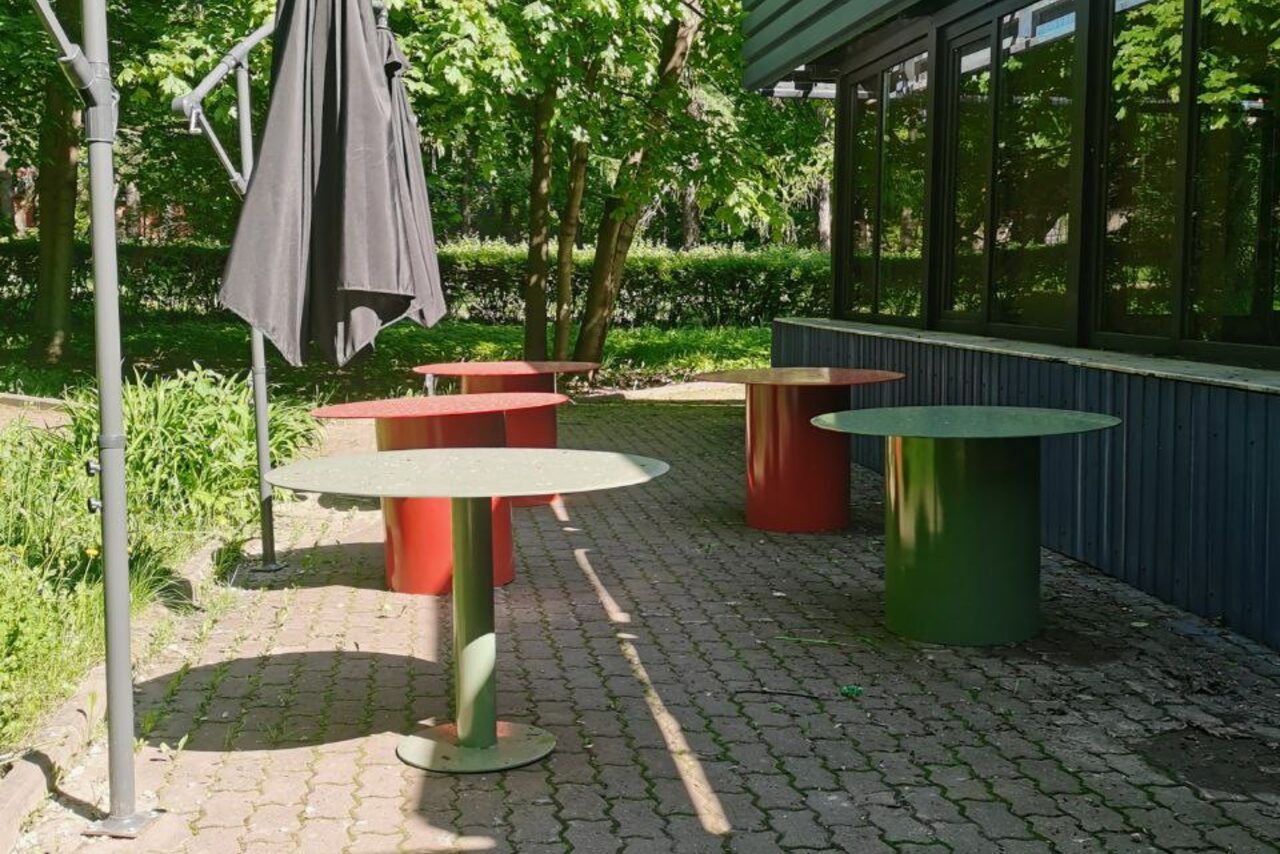 Арендовать Просторный и светлый лофт с панорамным видом в парк. Открытая площадка с зеленым и красным круглыми столами на мощеной поверхности. Черный закрытый зонтик. Зелень вокруг, окна сбоку. Тенистое освещение, солнечные пятна.