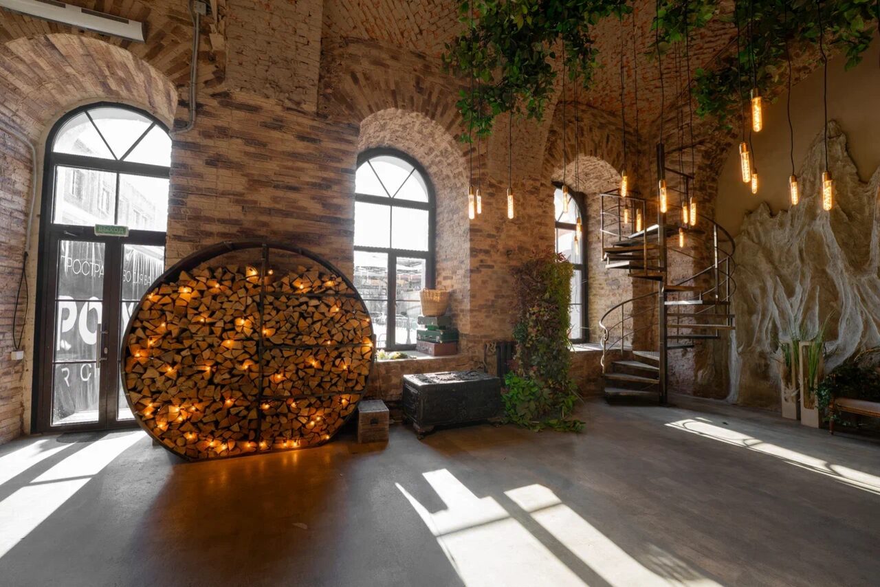 Арендовать Polygon(Полигон). Эко-Лофт. Просторное помещение с кирпичными стенами и большими арочными окнами. В центре круглая поленница с подсветкой. Спиральная лестница из металла. Потолок украшен свисающими лампами. На полу темное покрытие. Живые растения в кадре.