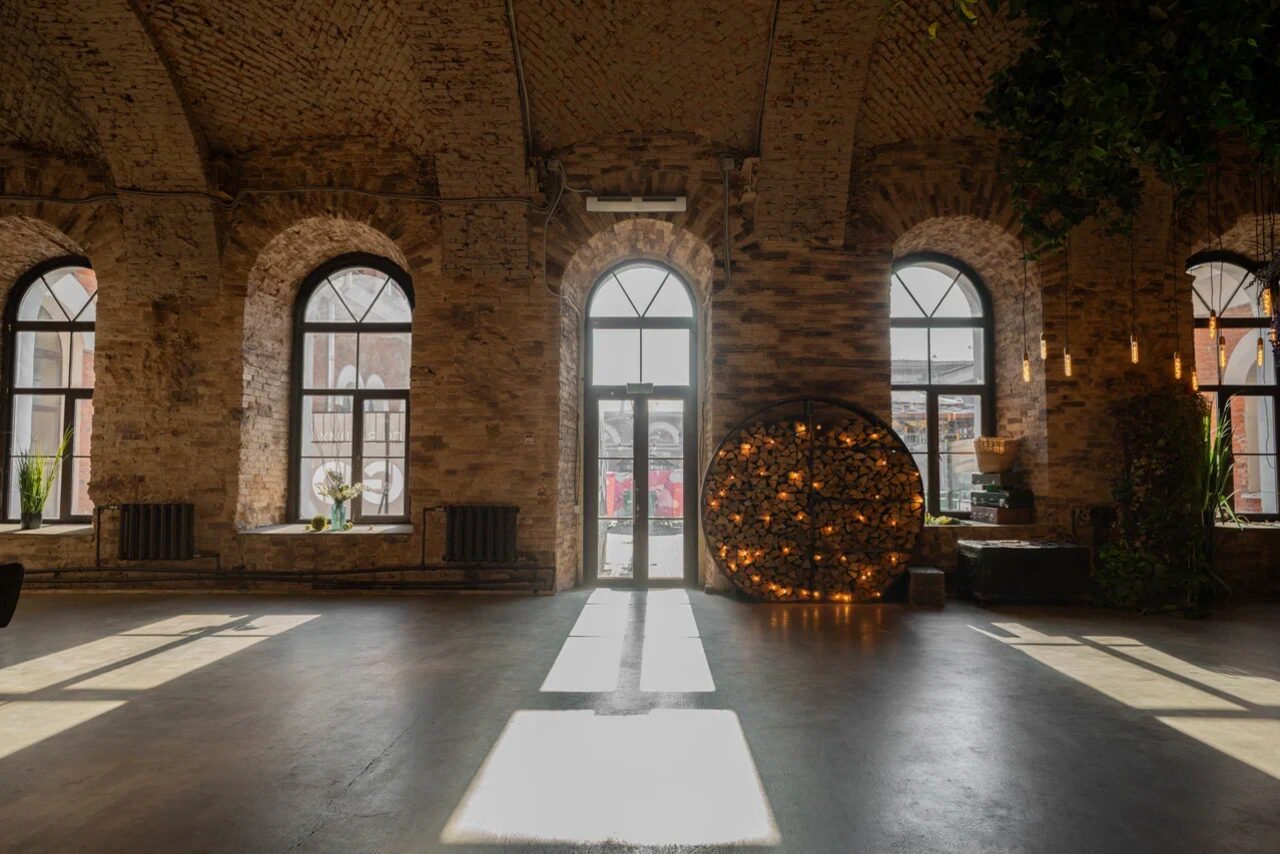 Арендовать Polygon(Полигон). Эко-Лофт. Просторное помещение с кирпичными стенами и сводчатым потолком. Множество высоких арочных окон дают естественное освещение. На полу серое покрытие. В углу круглая декорация с огоньками и комнатные растения. Потолочные лампы добавляют света.