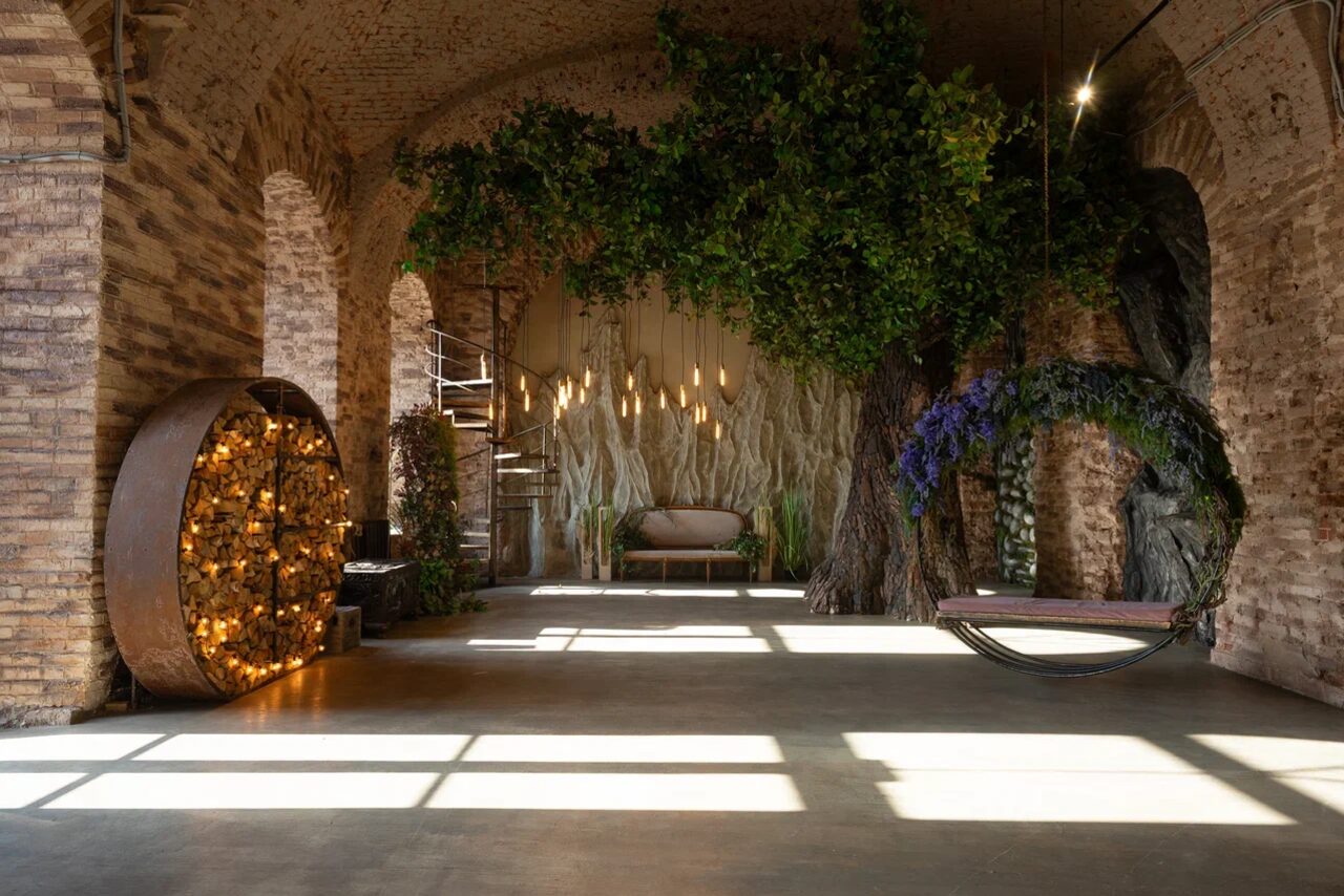 Арендовать Polygon(Полигон). Эко-Лофт. Арочный зал с кирпичными стенами и деревянным потолком, большой круглый шкаф с декоративной подсветкой, центральный диван, качели с цветочной декорацией. Искусственное дерево с листвой, подвешенные светильники, яркое естественное освещение через большие окна.