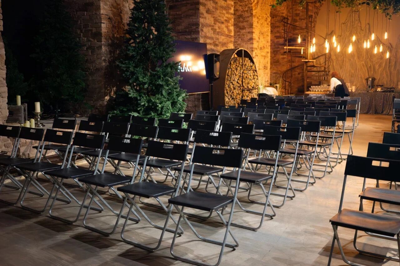 Арендовать Polygon(Полигон). Эко-Лофт. Пространство со складными стульями, оформленное в индустриальном стиле. Кирпичные стены, деревянный пол, декоративные деревья. На заднем плане стол, лампы Эдисона и витая лестница. Свет теплый, атмосфера уютная.