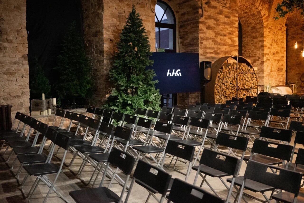 Арендовать Polygon(Полигон). Эко-Лофт. Просторное помещение с кирпичными стенами и арочными окнами. Ряды черных складных стульев. Большая елка рядом с экраном с белым логотипом на синем фоне. Декор из дров и приглушенное освещение.