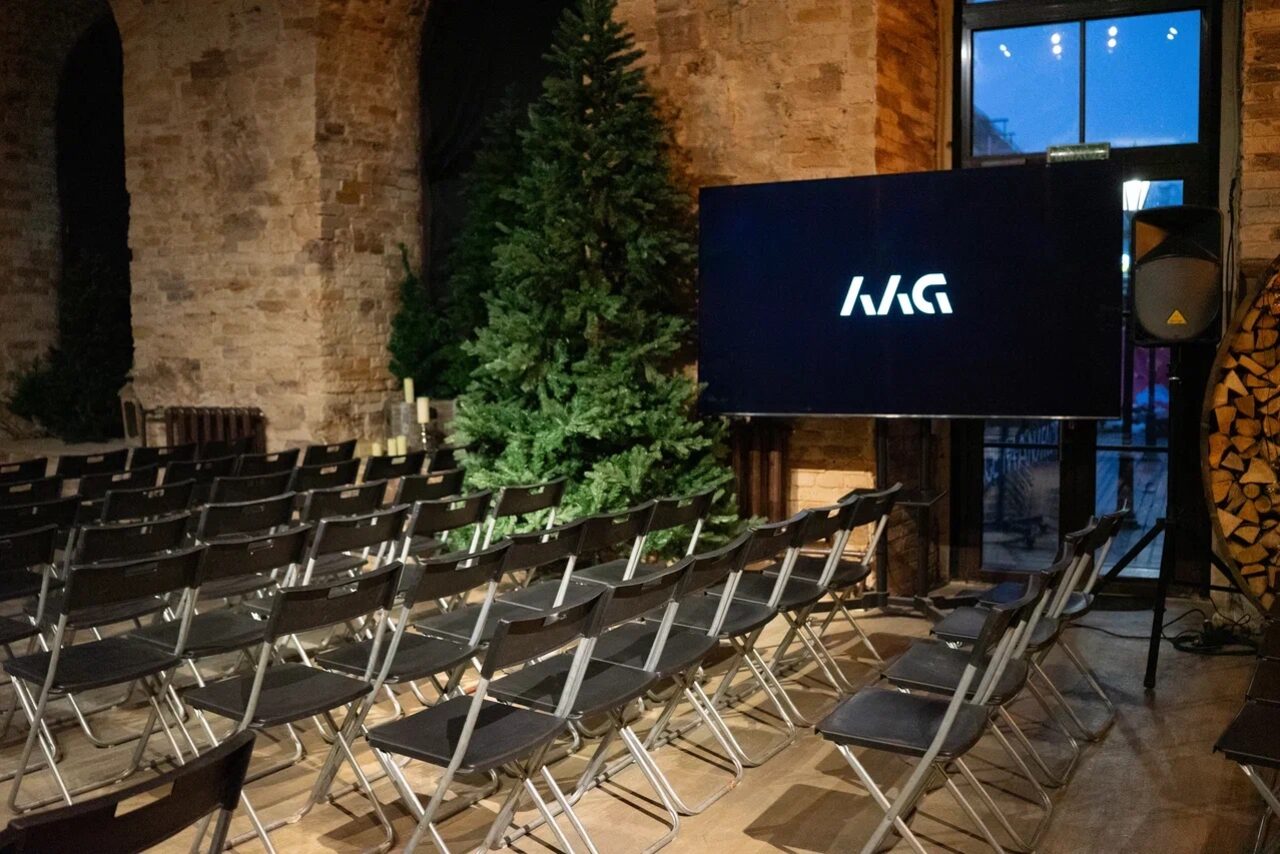 Арендовать Polygon(Полигон). Эко-Лофт. Аудиторное пространство с рядами черных складных стульев перед большим экраном с логотипом. Каменные стены, искусственная елка, стеклянная дверь. Деревянный пол, теплое освещение.