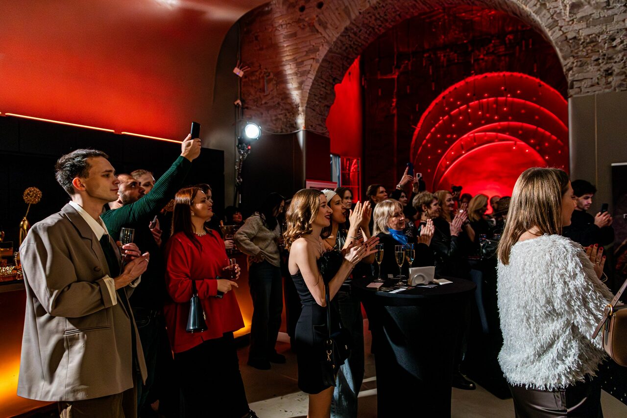 Арендовать Polygon(Полигон). Зал Кухня. Арочный кирпичный зал с приглушенным красным освещением и акцентными светильниками. В центре группа людей стоит вокруг высоких черных столов с напитками. Стены из камня, декорированы светодиодными полосами.