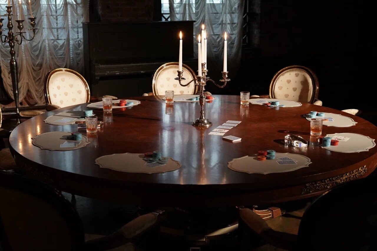 Арендовать Polygon(Полигон). Зал Апартаменты. Круглый деревянный стол с подсвечником на три свечи. Вокруг стола расположены стулья с мягкой обивкой. На столе разложены карточки, фишки и бокалы с напитками. Фон затемнён, видны свечи и занавески.
