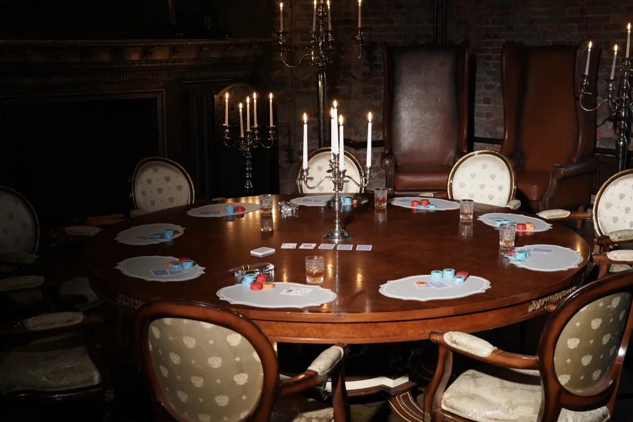 Арендовать Polygon(Полигон). Зал Апартаменты. Круглый деревянный стол с игральными картами и фишками, окруженный стульями с обивкой. На столе свечи в канделябрах. Темное помещение с кирпичными стенами и камином. Тусклое освещение от свечей.